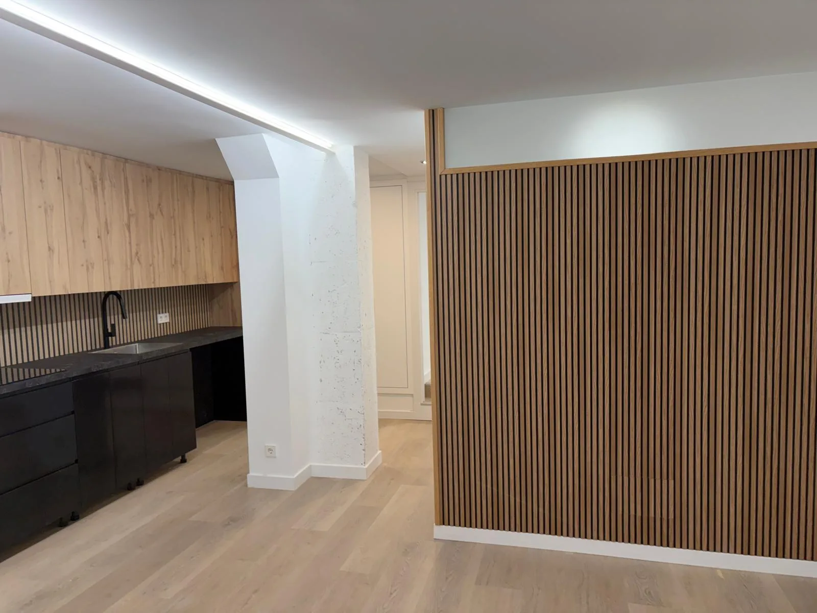 Espacio interior con suelo claro, cocina abierta y pared revestida con lamas verticales